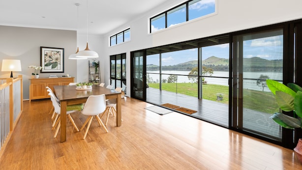 A modern kitchen with wooden cabinets, a dining table with chairs, and large windows offering a view of a lake and hills. - Bridge Water Views - Waterfront (Bonnie Doon)
