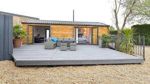 A modern outdoor living space with a wooden deck, a seating area with sofas and a table, and a bedroom with a bed and a nightstand. - Cheerful 1BD Cottage With Parking Nr Guildford (Guildford)