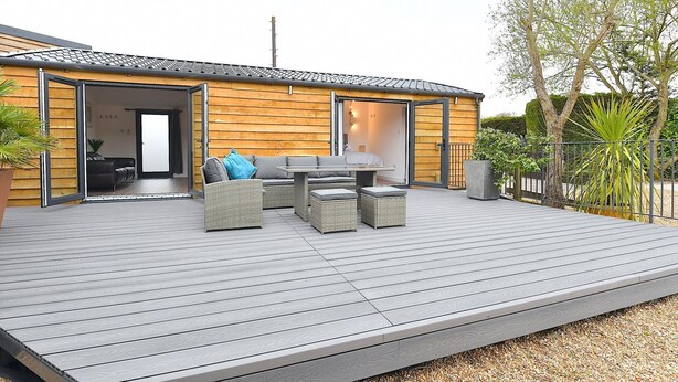 A modern outdoor deck with wicker furniture and a table, a wooden building with a sliding door, and a garden with a metal fence. - Cheerful 1BD Cottage With Parking Nr Guildford (Guildford)
