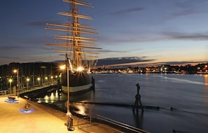 En strandpromenad med ett stort segelfartyg förtöjt vid kajen, en staty och en stadsbild i bakgrunden.
