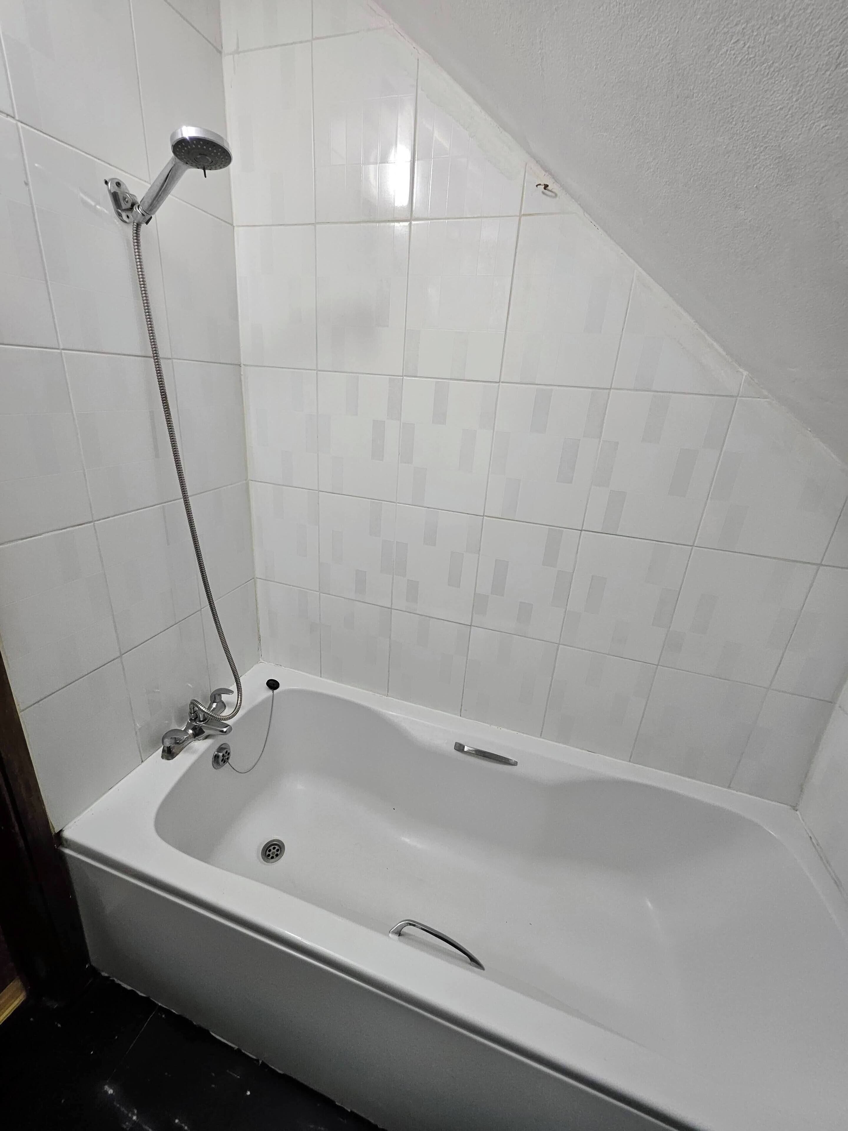 A standard rectangular bathtub with a handheld showerhead, a built-in drain, and a faucet. The room has white tiled walls and a sloped ceiling.