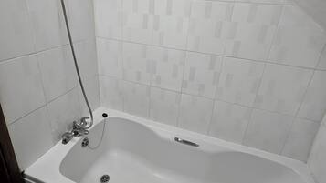 A standard rectangular bathtub with a handheld showerhead, a built-in drain, and a faucet. The room has white tiled walls and a sloped ceiling.