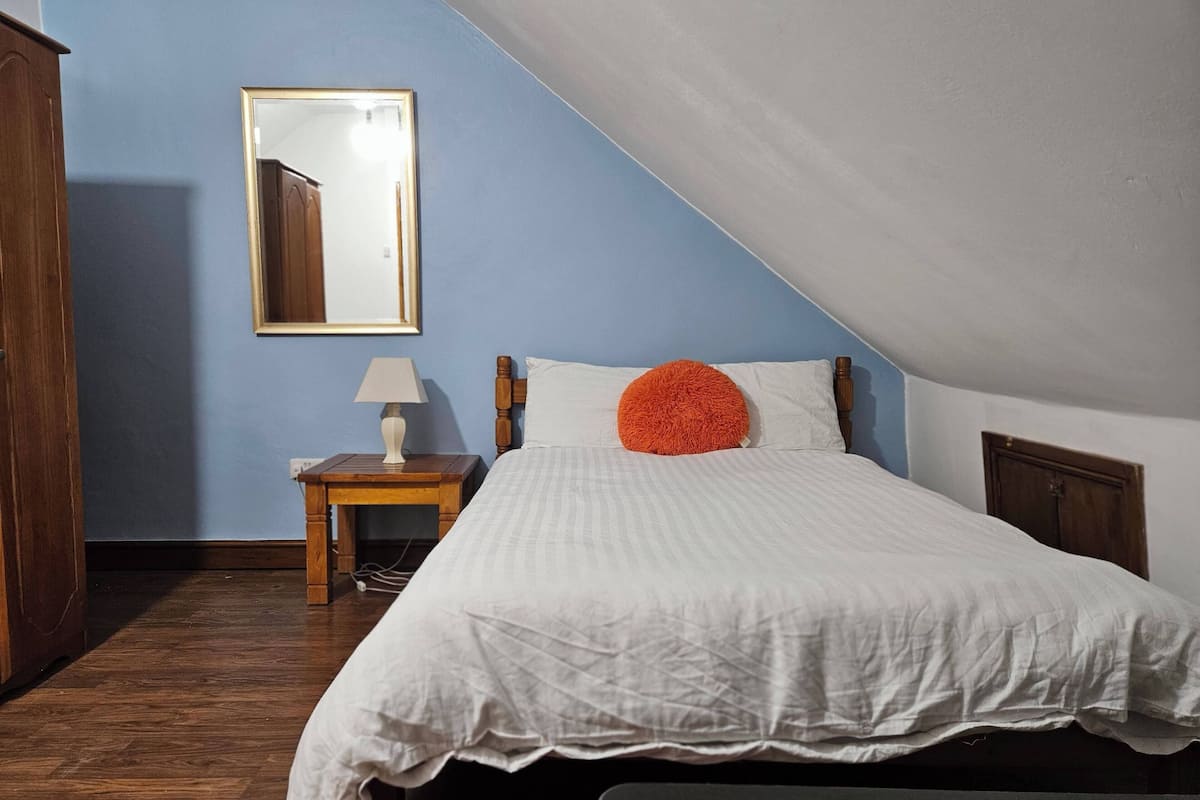Quarto com cama, mesa de cabeceira com abajur, espelho na parede e guarda-roupa de madeira.