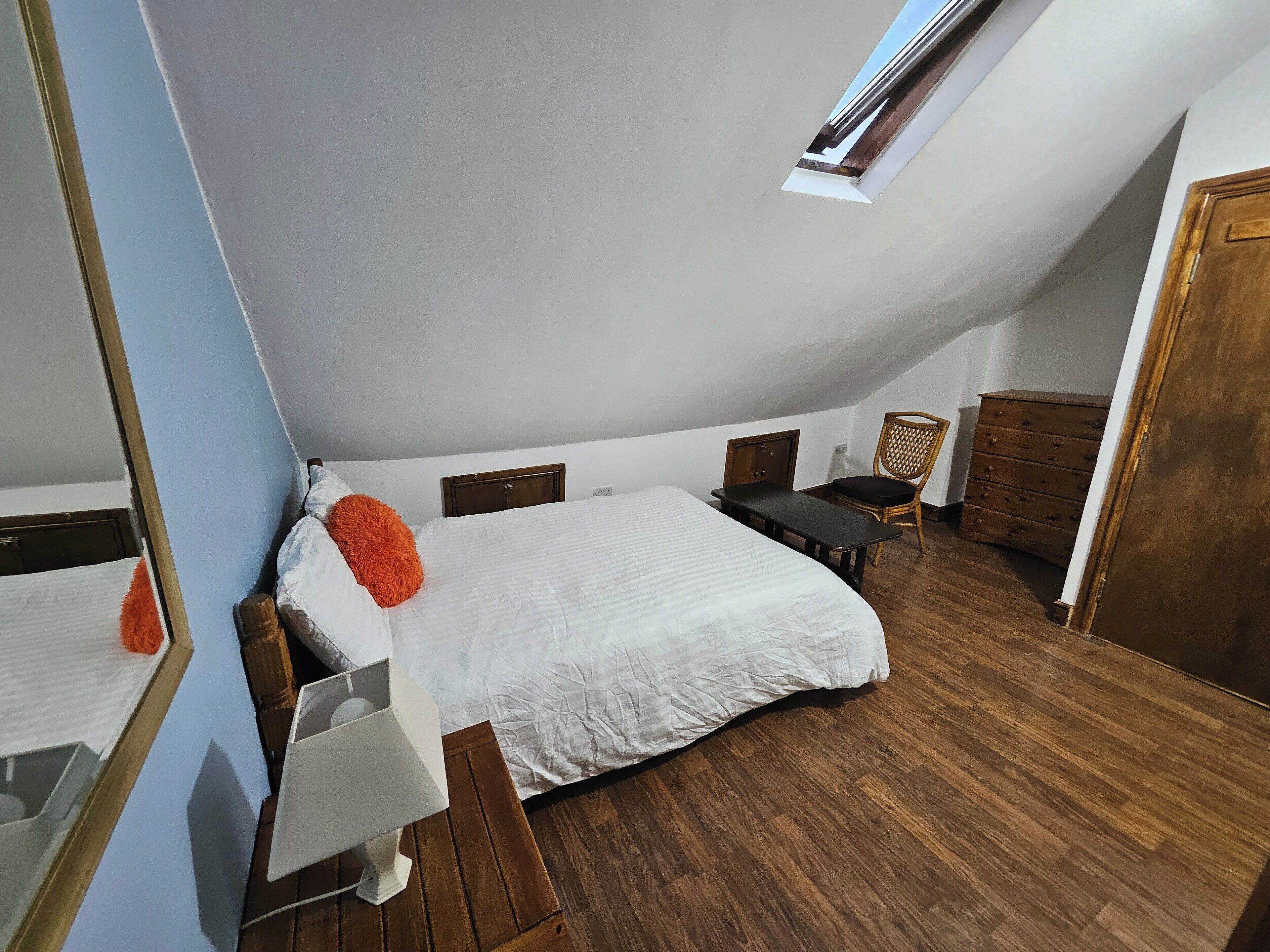 Un dormitorio con techo inclinado, una cama con ropa de cama blanca, un aparador de madera, una silla y una mesita.