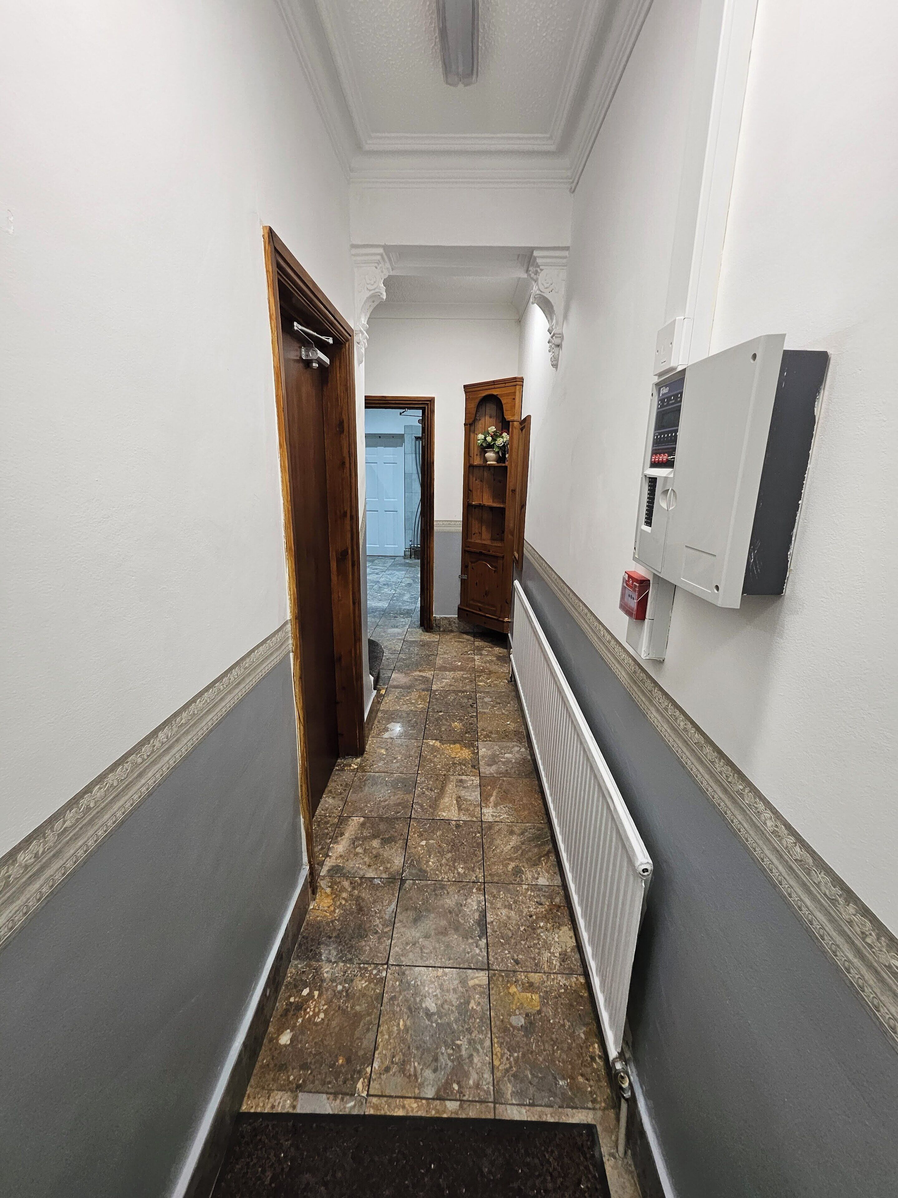 A hallway with a tiled floor, a radiator, a wooden door, and a wall-mounted electrical box.