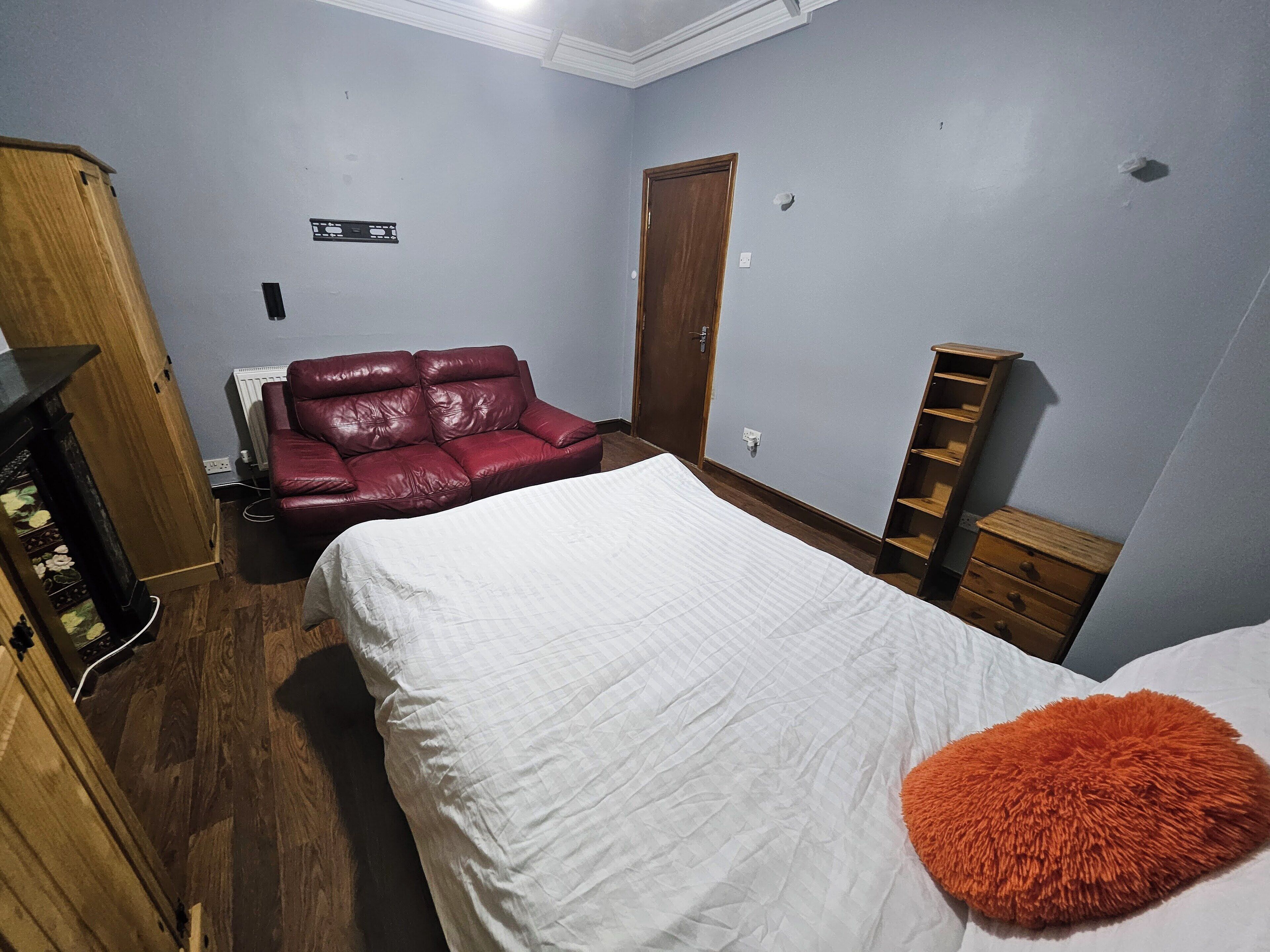 A bedroom with a bed, a red leather sofa, a wooden wardrobe, and a small wooden dresser.