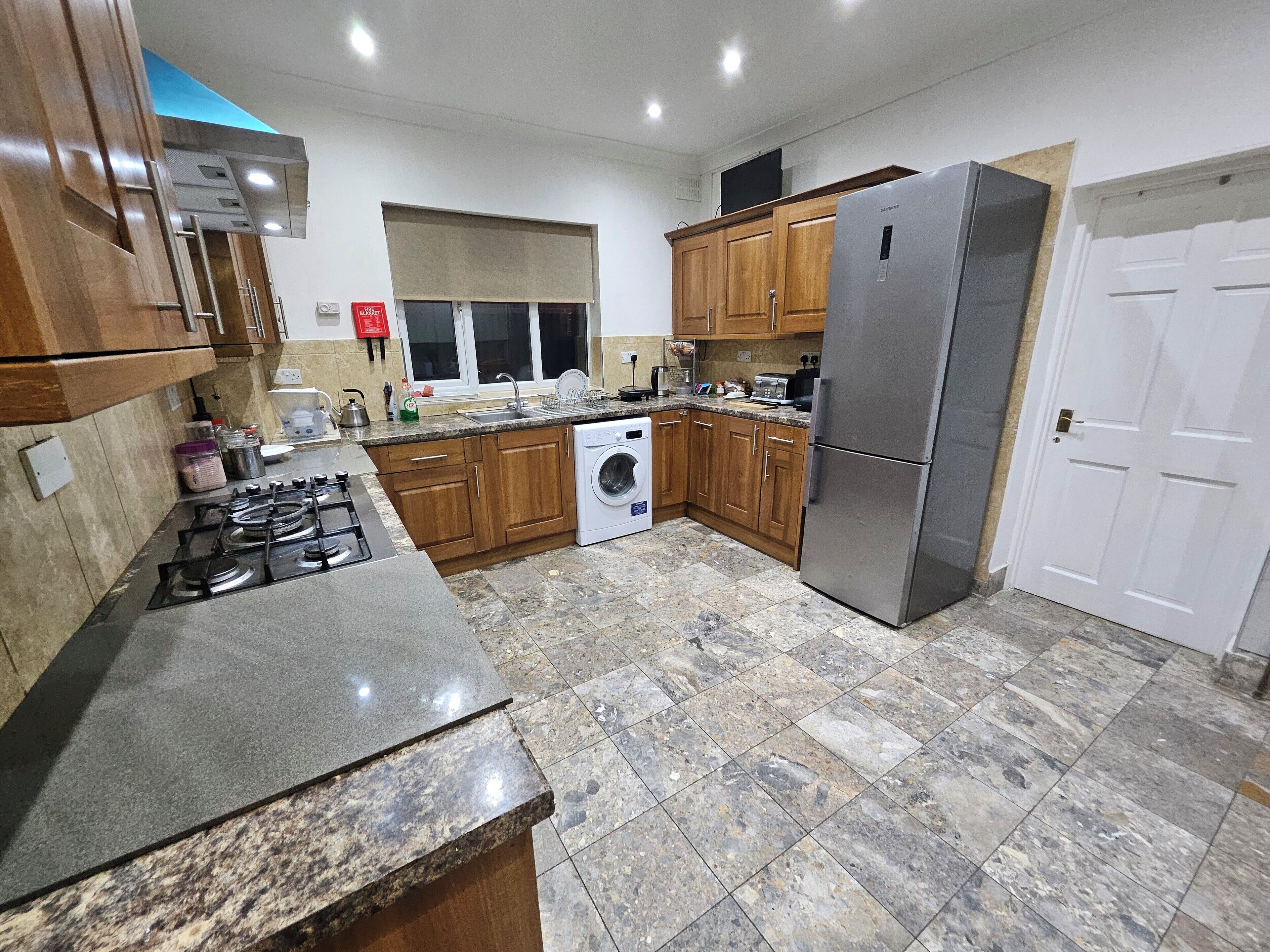 A kitchen with granite countertops, wooden cabinets, a stainless steel refrigerator, a washing machine, and a gas stove.