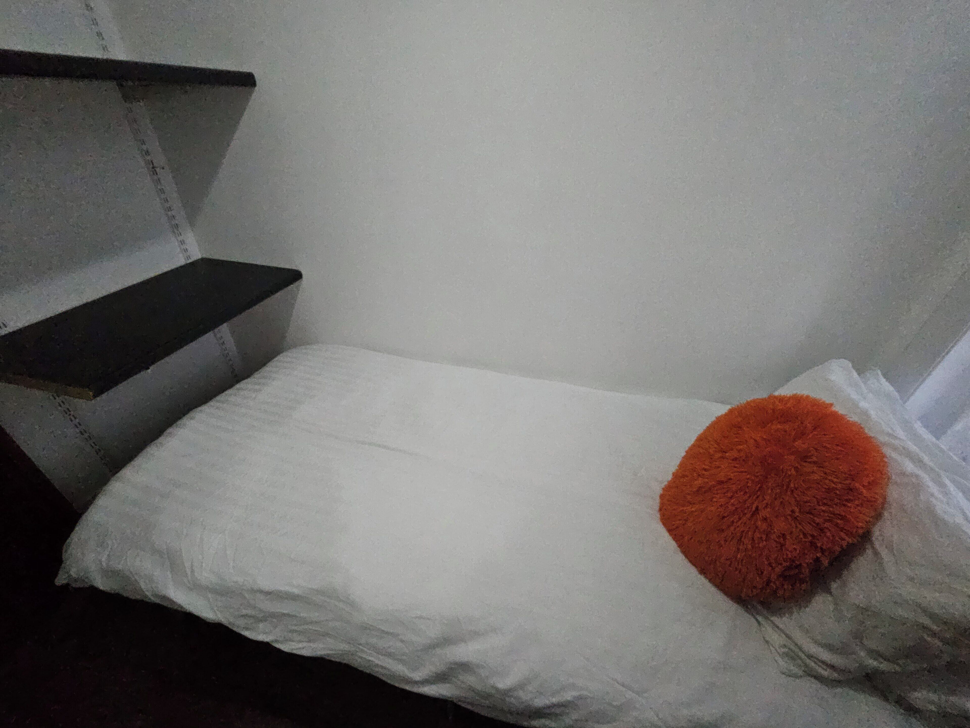 Uma cama pequena e bem arrumada com roupa de cama branca e um único travesseiro laranja.