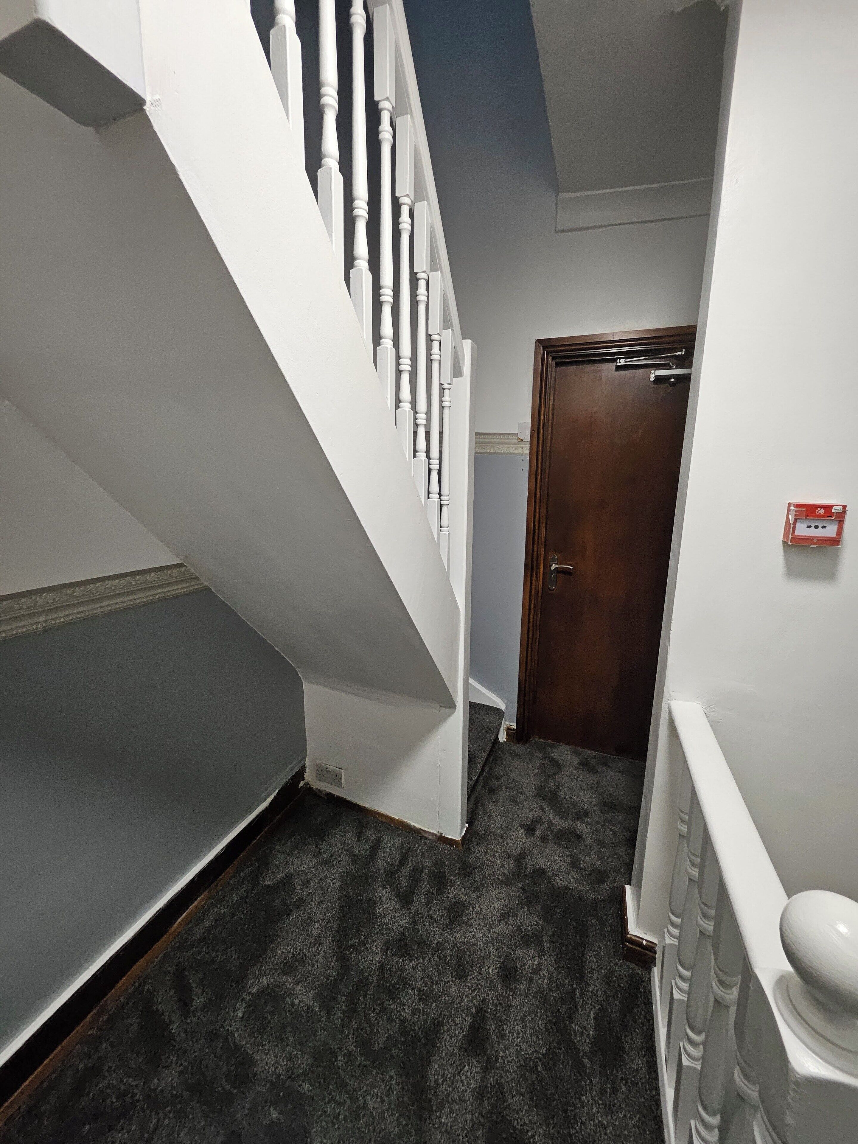 A narrow hallway with a carpeted floor, a wooden door, and a white railing.