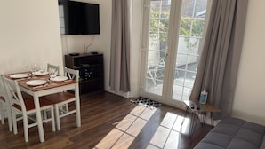 Een moderne woonkamer met een houten eettafel voor vier personen, een grijze bank, een aan de muur bevestigde televisie en een schuifdeur van glas naar een buitenruimte.