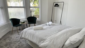 Een slaapkamer met een groot bed, twee stoelen, een vloerlamp en uitzicht naar buiten door de ramen.