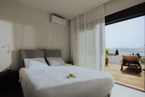 A modern bedroom with a large bed, a nightstand, an air conditioning unit, and a view of a balcony with a wooden deck and a chair. - Villa Pelagia Summary (Paleros)