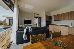 Un espacio habitable moderno con un sofá negro, una mesa de comedor de madera y una zona junto a la piscina.
