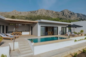 A modern outdoor pool area with a mountain backdrop, featuring a dining area and lounge chairs. - Villa Pelagia Summary (Paleros)