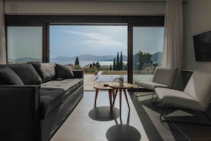 A modern living room with a sofa, armchair, and a small table, large windows with a view of the sea and mountains, and a flat-screen TV mounted on the wall. - Villa Pelagia Summary (Paleros)