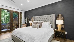 Une chambre avec un grand lit, des murs sombres et une vue sur une terrasse en bois et des arbres.