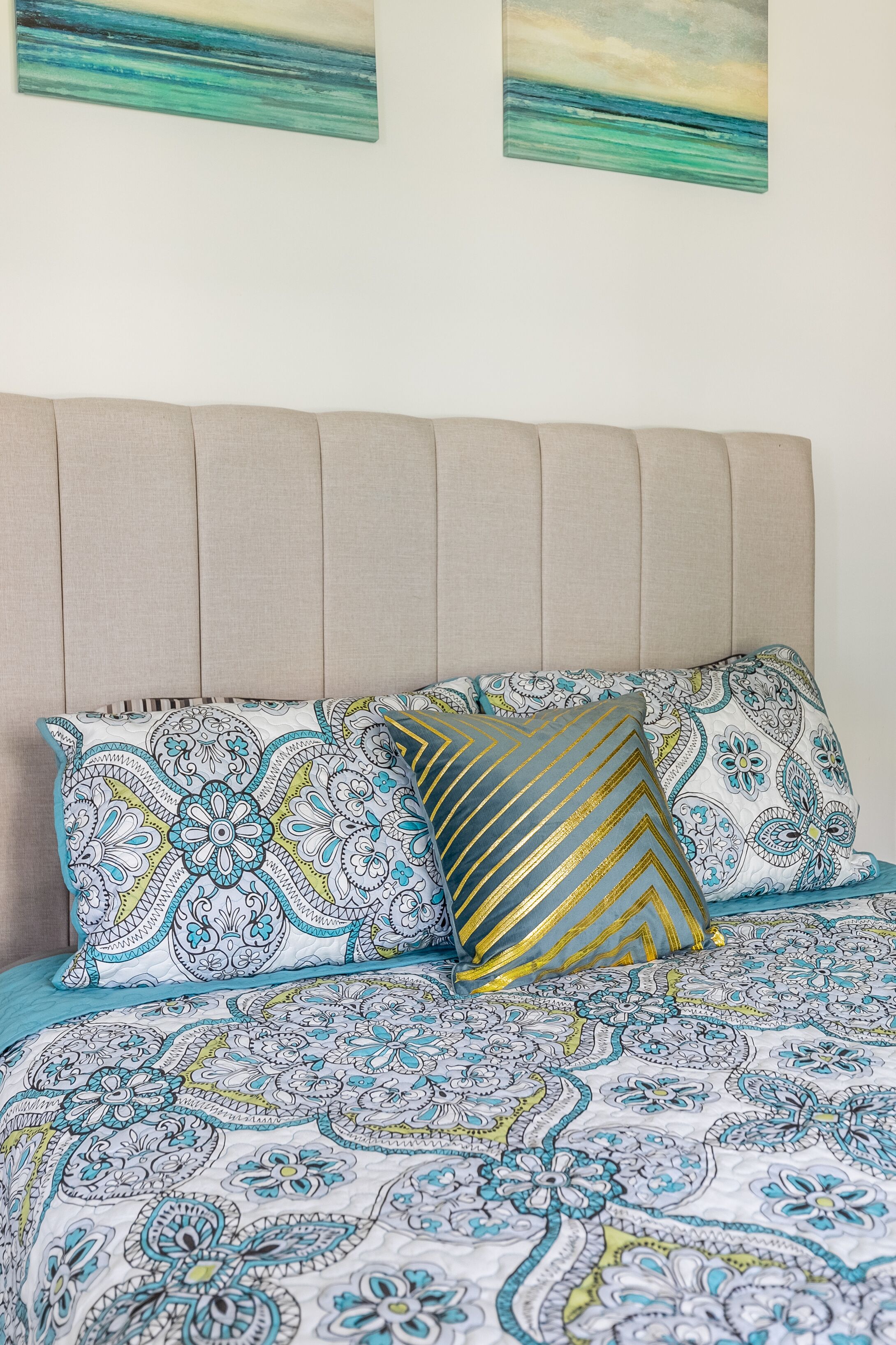 Una cama con ropa de cama estampada y almohadones decorativos, un cabecero capitonado y dos obras de arte abstracto en la pared.