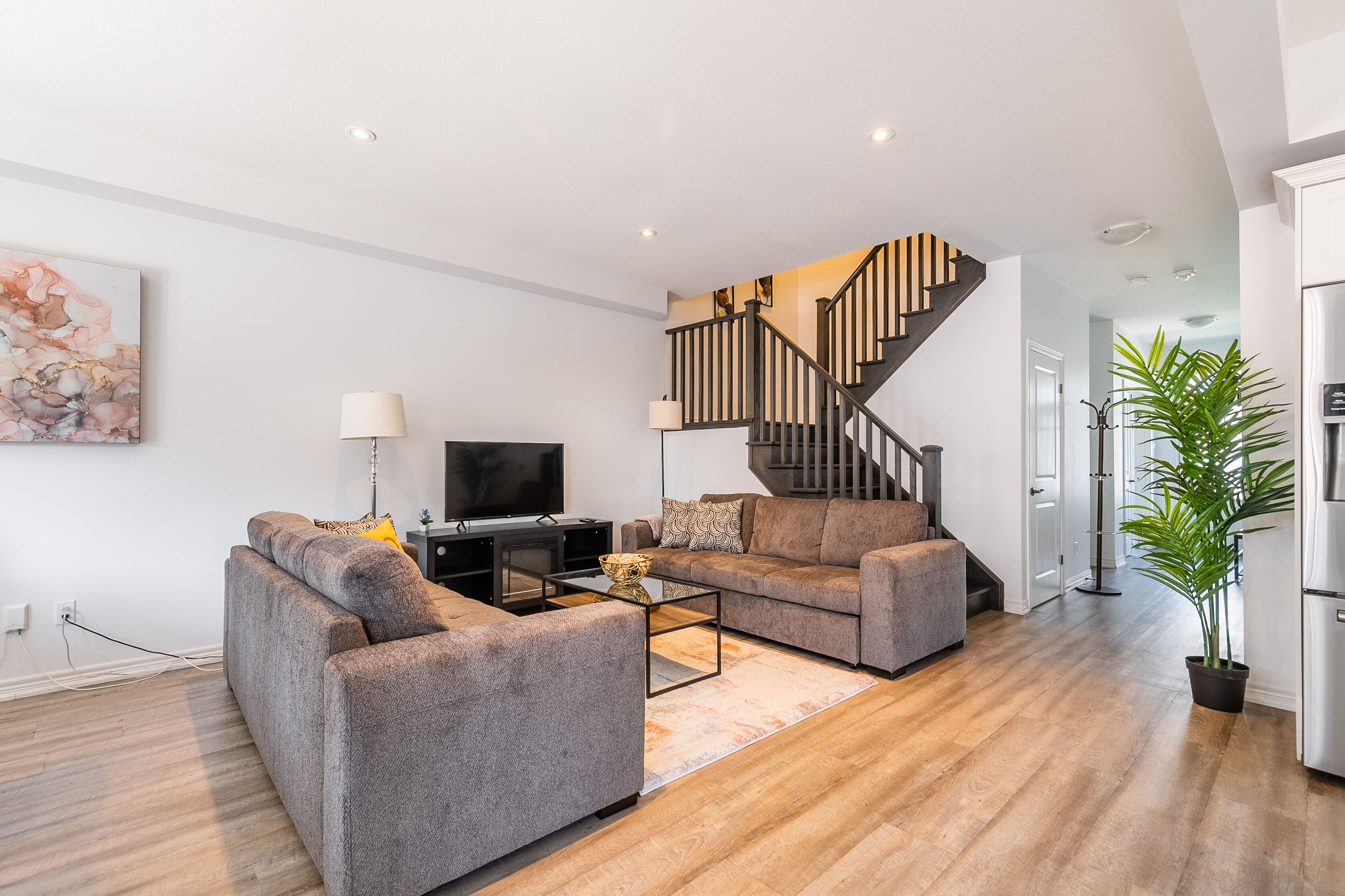 A modern living room with a sofa set, a coffee table, and a TV. A staircase with a wooden railing leads to an upper floor.
