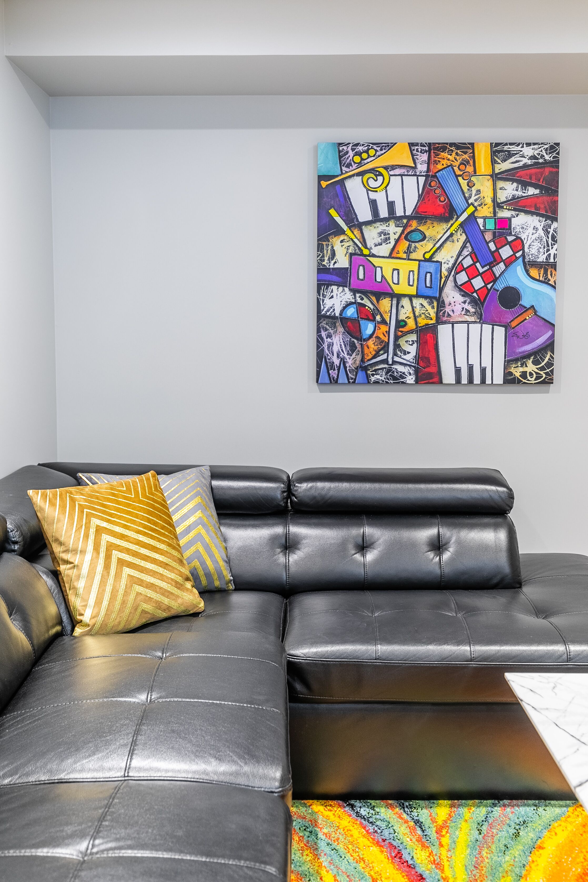 Un canapé d’angle en cuir noir agrémenté de deux coussins à motifs, un tableau abstrait aux couleurs vives accroché au mur et un plaid multicolore posé sur le sol.