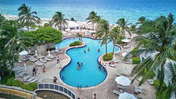 A resort pool with a view of the ocean, surrounded by palm trees and lounge chairs.