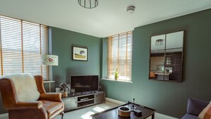 A living room with a leather armchair, a sofa, a coffee table, a TV, and a kitchen area visible through a window. - Elegant & stylish home in historic town centre - Pass the Keys (Moreton-in-Marsh)