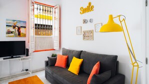 Een woonkamer met een grijze bank, oranje en gele kussens, een gele vloerlamp, een televisie op een witte dressoir, en een schilderij aan de muur.