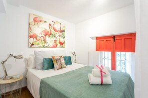 A bedroom with a bed, pillows, a desk, a lamp, a painting, and a window with a red shutter.