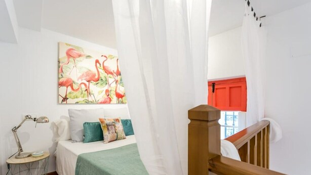 Een slaapkamer met een bed, een bedlamp, een schilderij, een raam met rood gordijn en een houten leuning.
