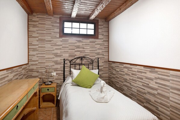 A small, narrow room with a bed, a wooden nightstand, a window, and a tiled wall.