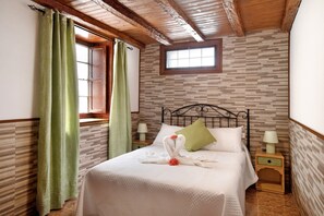 Una cama bien tendida con un almohadón verde, dos mesitas de noche con lámparas, una ventana con cortinas y un papel tapiz estampado.