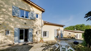 Un bâtiment à deux étages avec une allée en gravier, un espace extérieur aménagé pour s’asseoir et un barbecue.