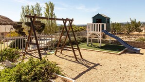 Un parque infantil con columpios, tobogán y estructura para escalar, rodeado por una valla y follaje.