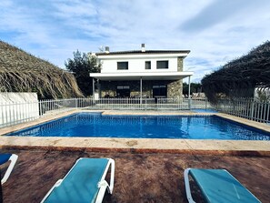A swimming pool with lounge chairs, a white fence, and a house with a stone facade. - Villa Casa Rural la Montana 2 with Pool, Garden & Wi-Fi (Chella)