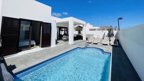 Una piscina con borde de azulejos, un área de estar techada con sillas y un edificio blanco con puerta oscura.