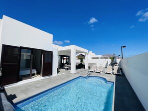 Una piscina con borde de azulejos, un área de estar techada con sillas y un edificio blanco con puerta oscura.