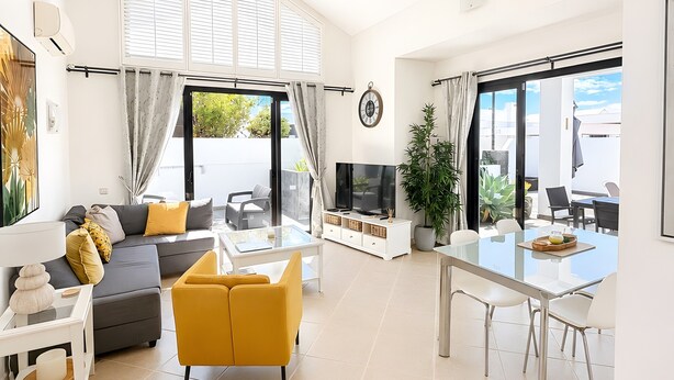Una sala de estar moderna con sofá, sillón amarillo, televisor, mesa de comedor y ventanas grandes.