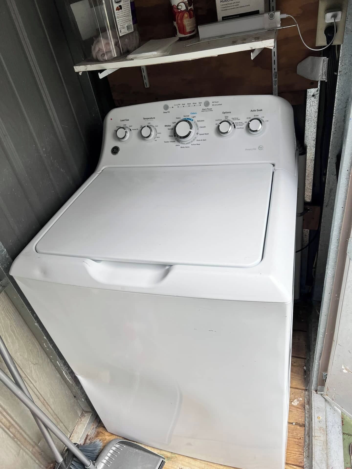 A compact, white front-loading washing machine in a small utility room with a metal shelf above it.
