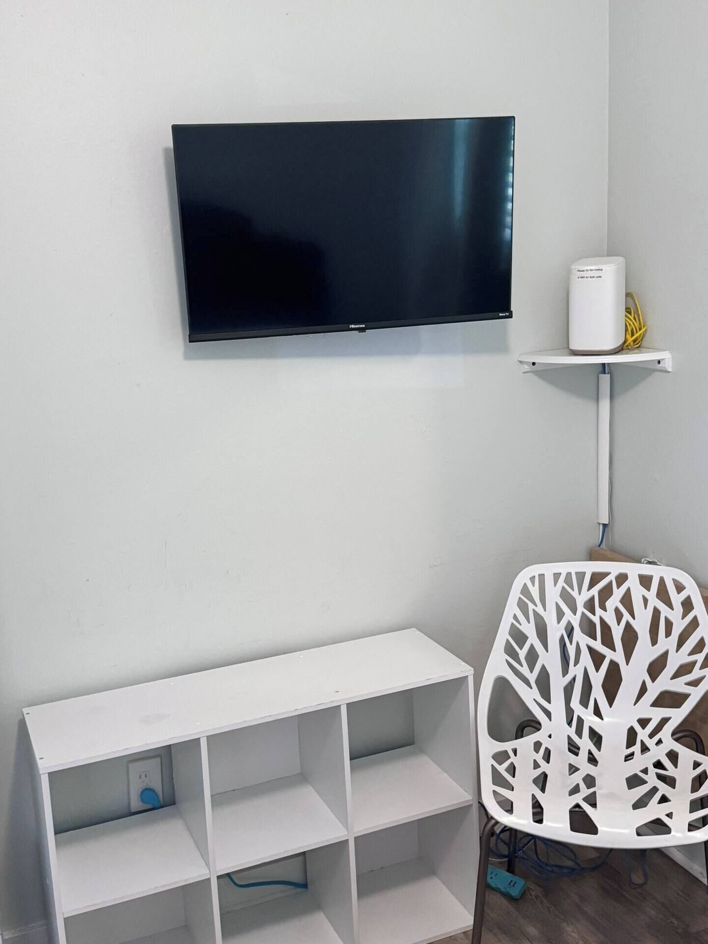 A room with a flat-screen TV mounted on the wall, a white shelf with a device, a white chair with a tree-like pattern, and a white shelving unit with multiple compartments.