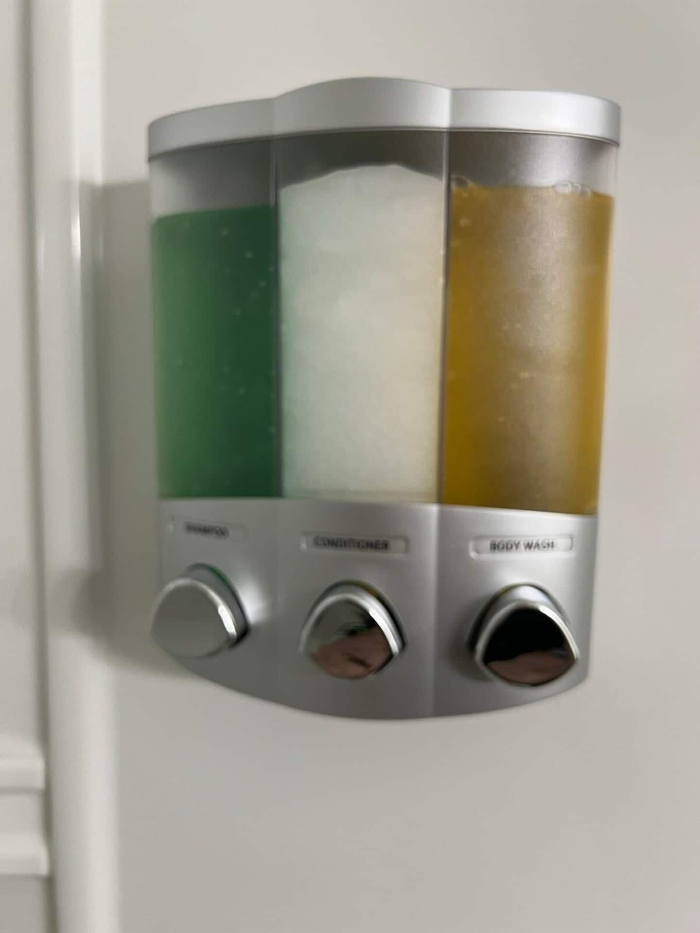 A wall-mounted dispenser with three compartments for shampoo, conditioner, and body wash.