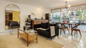 A modern living room with a sofa, coffee table, and dining area. A kitchen is visible in the background, and there is a large window providing a view of the outdoors. - w* | Splendid 1BR with Perfect Balcony in Polanco (Ciudad de México)