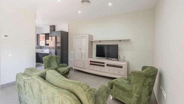 A living room with a flat-screen TV, green sofas, a cabinet, and a kitchen in the background. - Holiday Apartment 'Torero II' with Pool, Private Balcony and Wi-Fi (Sevilla)