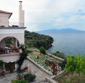 Una propiedad costera con patio de piedra, mobiliario exterior y vistas al mar y a las montañas.