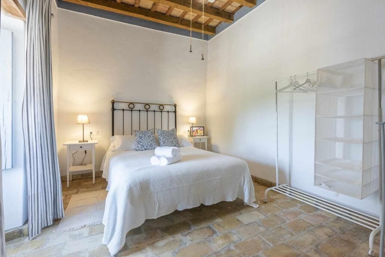 Un dormitori amb sòl de pedra, un llit amb roba de llit blanca, dues làmpades de nit, una estant per a la roba penjada i una finestra amb cortines.