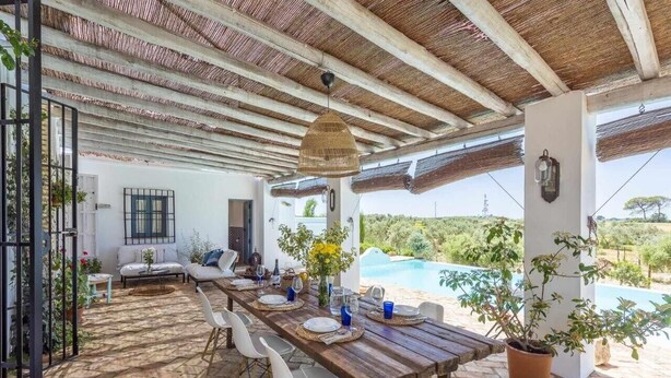 Ein Essbereich im Freien mit einem Holztisch, gedeckt für vier Personen, umgeben von weißen Stühlen, einer hängenden Lampe und Blick auf Pool und Grünflächen.