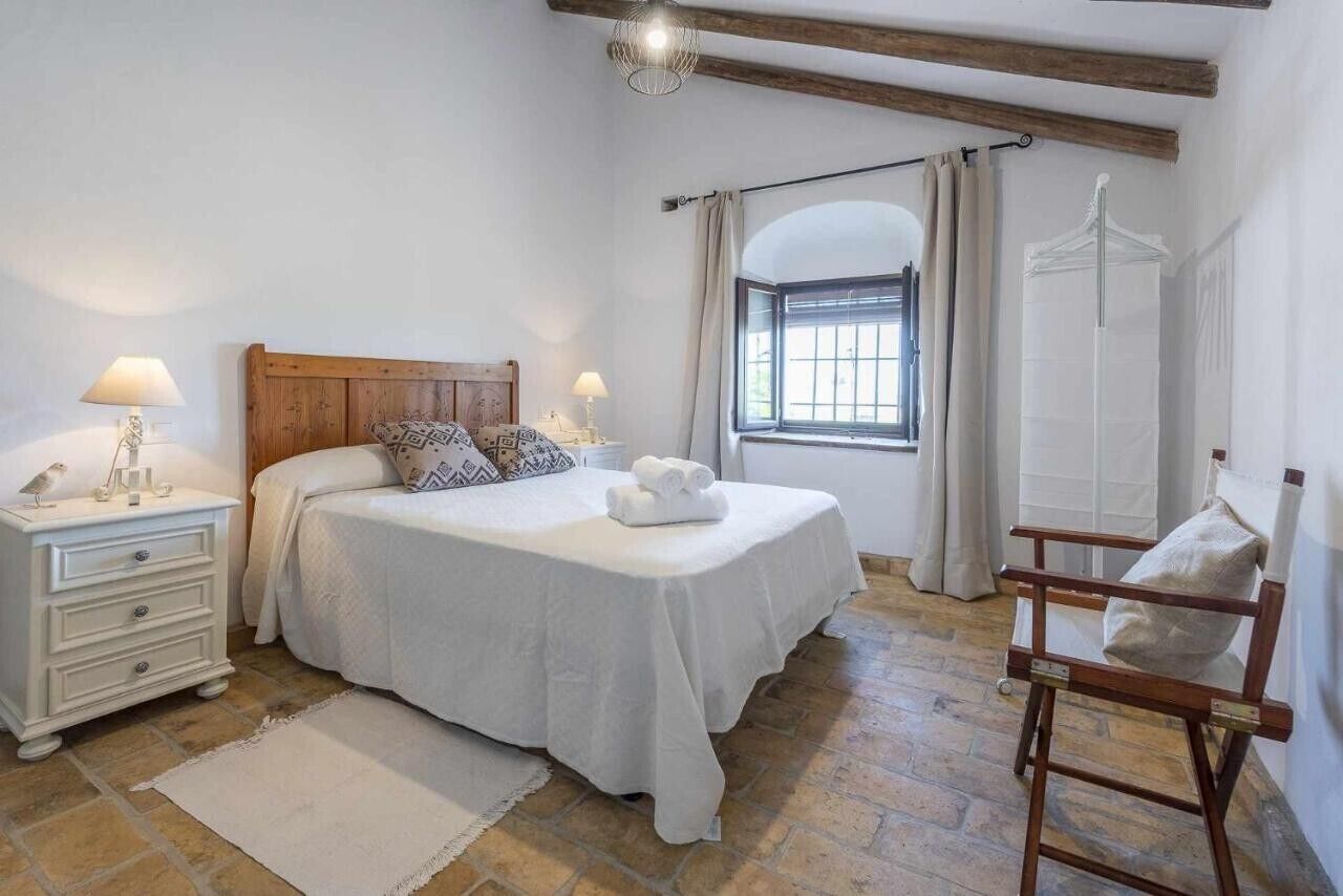 Un dormitorio con cama, mesitas de noche, una silla, un ventanal y un armario.