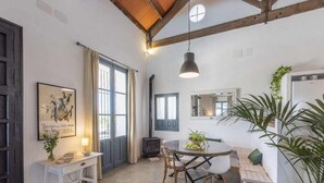 Ein gemütliches Zimmer mit Holzboden, einem Esstisch mit Stühlen, einem Sofa, einem kleinen Tisch mit Lampe, einer Topfpflanze und einem großen Fenster mit Vorhängen.