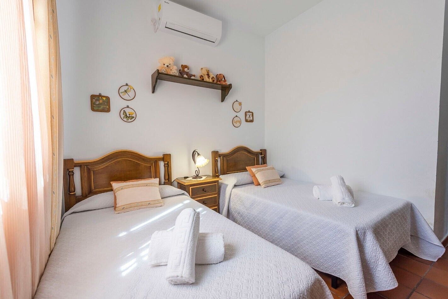 Une chambre d’hôtel avec deux lits, une table de chevet avec une lampe, une étagère avec des objets décoratifs et un appareil de climatisation.