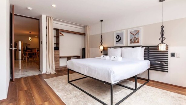 Une chambre moderne avec un lit, un plancher en bois, un tapis blanc et un coin repas visible à travers une porte ouverte.