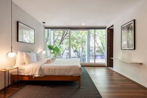 Une chambre à coucher moderne avec un grand lit, une vue sur les arbres par une porte coulissante et des œuvres d’art encadrées accrochées aux murs.