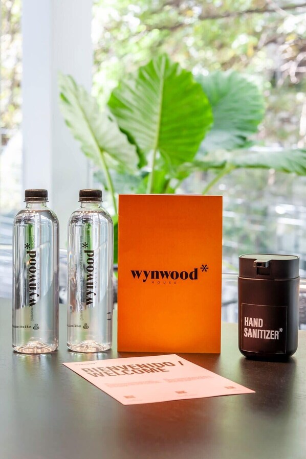 Une table avec deux bouteilles en verre étiquetées « Poxum », une boîte orange avec l’inscription « wynwood HOUSE » et un récipient noir étiqueté « GEL HYDROALCOOLIQUE ».